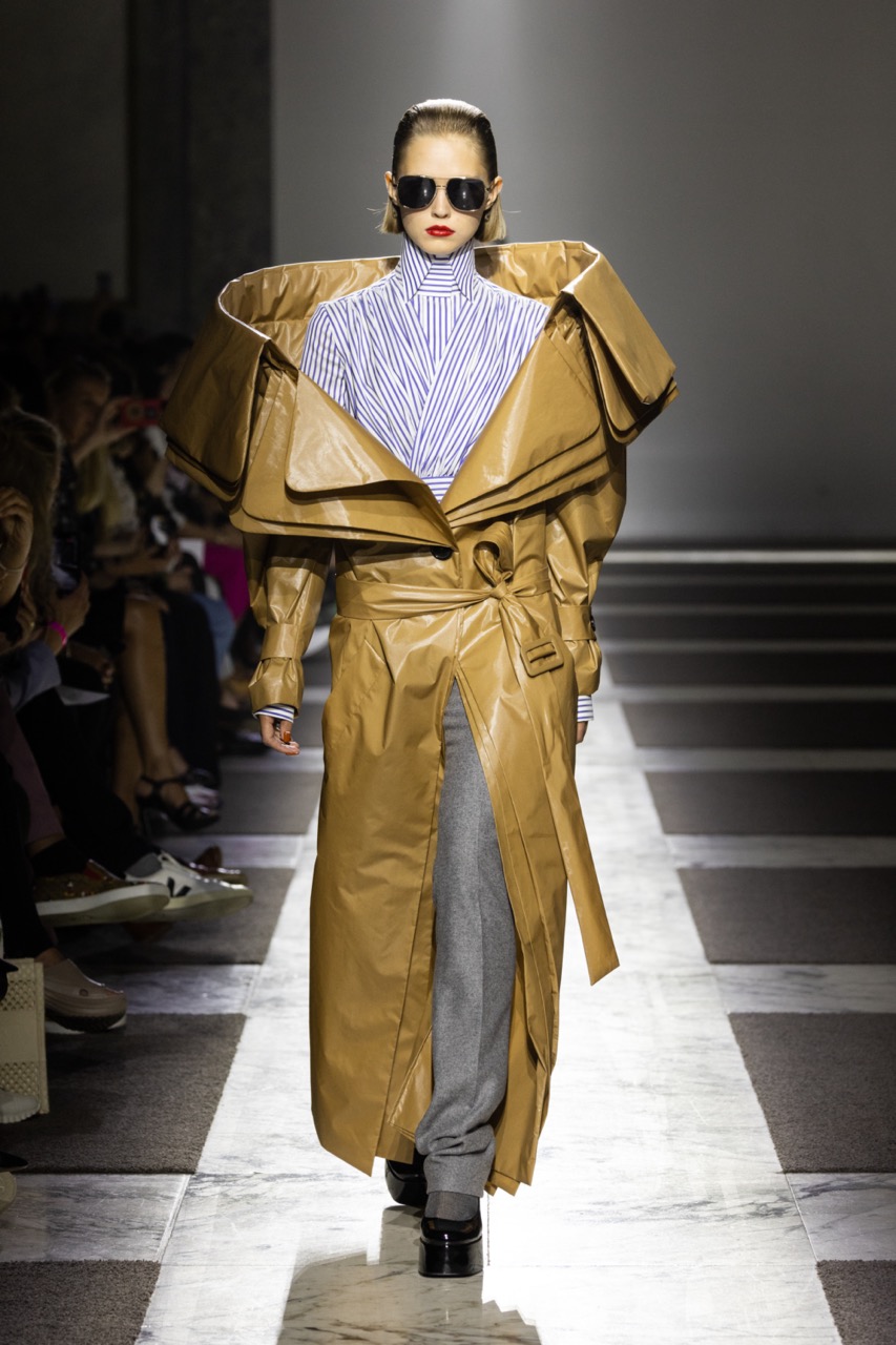  A draped, high-collared silk blouse is paired with a coated trench coat featuring a sculpturally layered collar and triple facing. The design is matched with grey trousers crafted from wool.